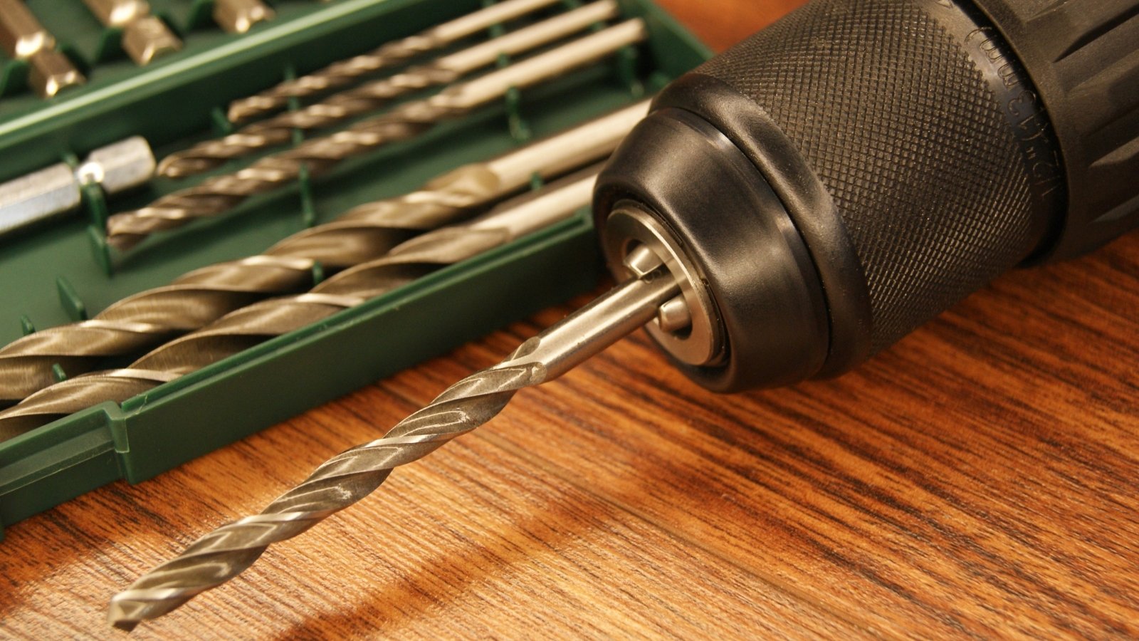 A black handheld drill with several shiny drill bits placed beside it on a wooden surface, ready for use.