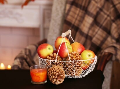 On a black serving table, a white basket overflows with apples and pinecones. Beside it, a solitary pinecone rests near a glowing candle in a glass holder. The white room's festive ambiance includes a beautifully decorated cloth.