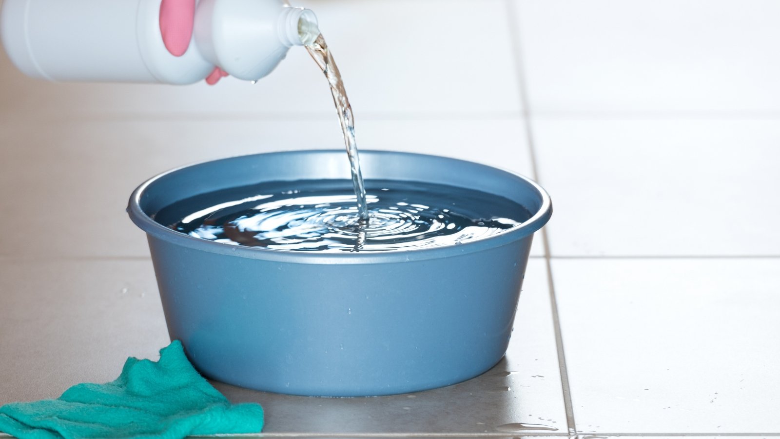 A light blue basin being filled with water from a white bottle, with a green cleaning cloth placed beside it on the tiled floor.