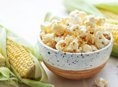 Grow popcorn. Close-up of a white ceramic bowl with ready-made popcorn on a white table next to three corn cobs. The corn cob, the reproductive structure of the corn plant, is characterized by its cylindrical shape and rows of kernels arranged around a central core. The kernels are pale yellow and glossy. The cob itself is encased in several layers of protective husks. Popcorn represents popped kernels are fluffy and irregularly shaped, with a crisp outer surface and a light, airy interior.