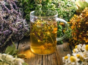 Close-up of a mug of brewed tea from herbal tea blends on a wooden table. A glass cup is filled with brewed green herbal tea with various dry herbs. Around the cup of tea there are bunches of dry herbs such as lavender, chamomile, linden and others.