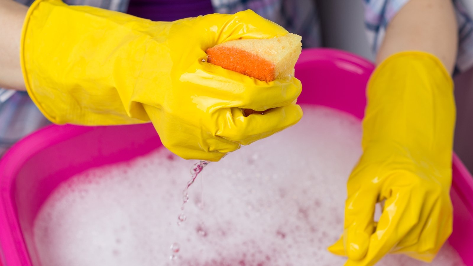A yellow-gloved hand holding an orange sponge above a pink tub filled with soapy water, with suds visible.