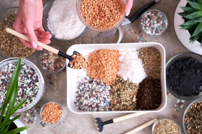 Preparing succulent soil substrate on a white table. On the table there is one large white plastic tray with piles of various components for preparing the substrate. Women's hands pour small orange pebbles from a glass bowl using a small spatula.